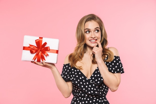 Una hermosa mujer joven y bonita posando aislada sobre la pared rosa sosteniendo la caja de regalo actual