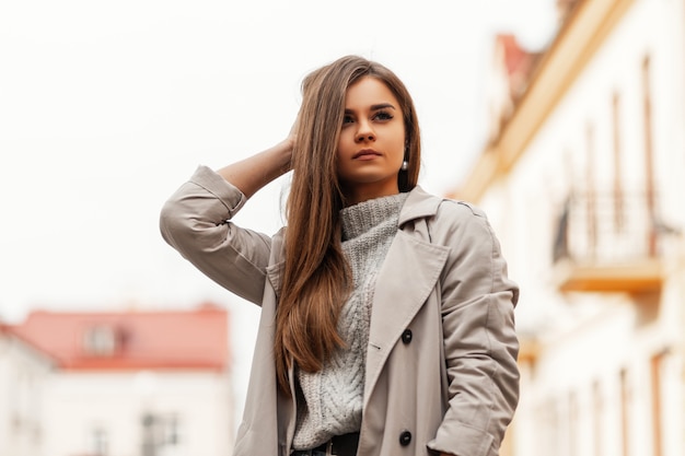 Hermosa mujer joven con cabello largo castaño en una elegante gabardina posando en la ciudad al aire libre