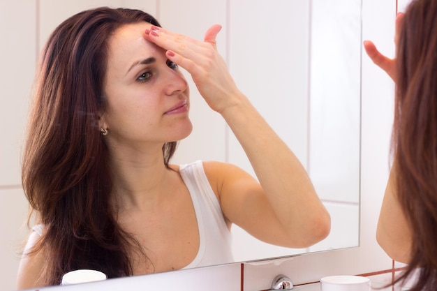 Hermosa mujer joven con cabello largo y oscuro en camisa blanca usando crema facial frente al espejo en su baño