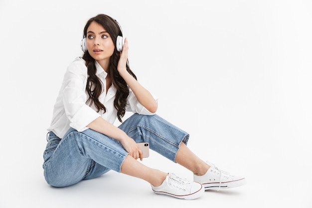 Hermosa mujer joven con cabello largo y rizado morena con camisa blanca sentado aislado sobre una pared blanca, disfrutando de escuchar música con auriculares