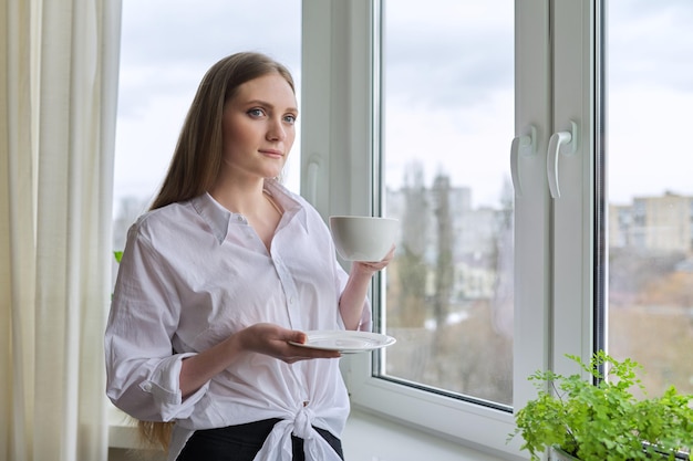 Hermosa mujer joven con cabello largo rubio sosteniendo una taza de café cerca de la ventana