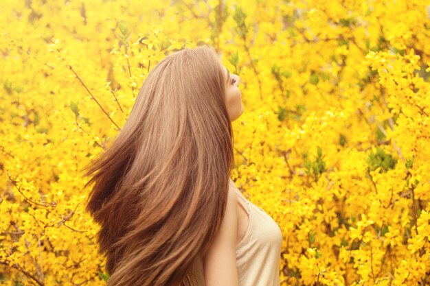 Hermosa mujer joven con cabello largo y saludable contra el fondo de flores amarillas Chica con retrato de peinado que sopla