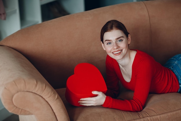 Hermosa mujer joven con cabello rojo acostado en su sofá en la sala de estar y sosteniendo la caja de regalo en forma de corazón y sonrisa Día de San Valentín o cumpleaños