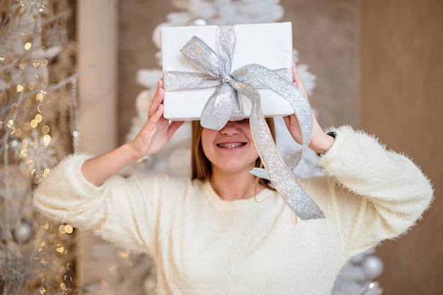 Una hermosa mujer joven con cabello rubio se encuentra cerca de un árbol de Navidad con un regalo en sus manos. El año nuevo llegará pronto. Ambiente navideño en un hogar acogedor.