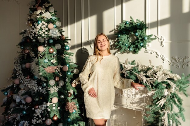 Una hermosa mujer joven con cabello rubio se encuentra en el contexto de un árbol de Navidad. El año nuevo llegará pronto. Ambiente navideño en un hogar acogedor.