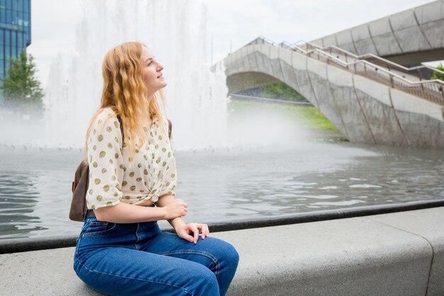 Hermosa mujer joven con cabello rubio de pie junto a la fuente. Chica relajarse en la calle en un día soleado. Concepto de viajes y vida activa. Al aire libre
