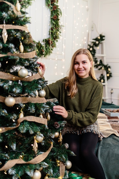 Una hermosa mujer joven con cabello rubio se sienta cerca de un árbol de Navidad y decora con juguetes. El año nuevo es pronto. Ambiente navideño en un hogar acogedor.