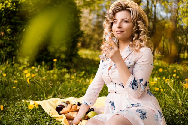 Hermosa mujer joven con cabello rubio en sombrero de paja bebe vino y sentado en el plaid en el jardín durante un picnic de verano