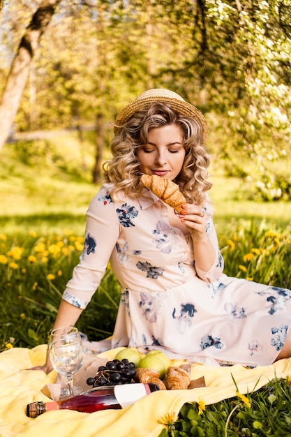 Hermosa mujer joven con cabello rubio con sombrero de paja y vestido blanco en el plaid en el parque. Mujer con un croissant en un picnic