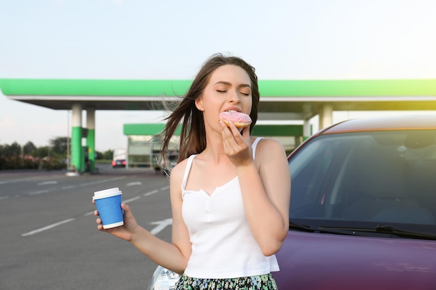 Hermosa mujer joven con café comiendo donut cerca del coche en la gasolinera