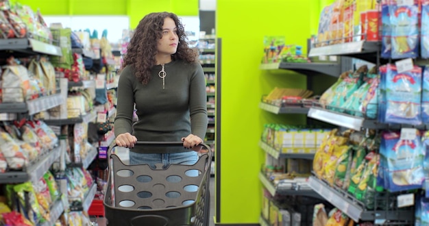 Hermosa mujer joven caminando con carrito de compras en el supermercado Joven mujer caucásica empujando carrito de compras con productos