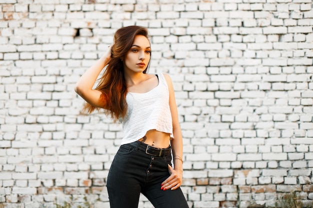 Hermosa mujer joven en una camiseta blanca y jeans negros cerca de una pared de ladrillo blanco.