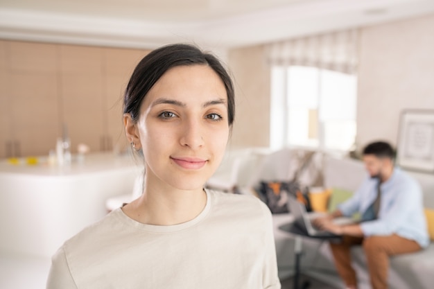 Hermosa mujer joven en camiseta blanca mirándote mientras está de pie frente a la cámara en la sala de estar contra su esposo usando una computadora portátil