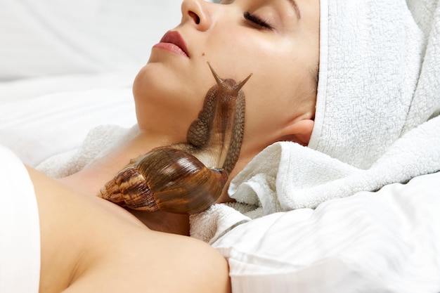 Hermosa mujer joven con un caracol en su rostro acostado en una cama blanca, procedimiento cosmético.