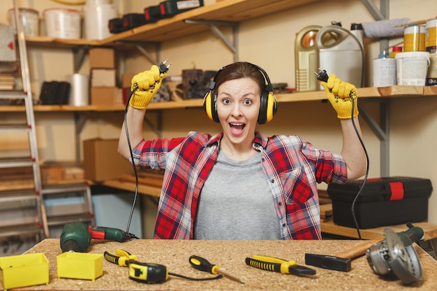 Hermosa mujer joven caucásica de diversión loca en camisa a cuadros, auriculares con aislamiento acústico que trabajan en el taller de carpintería en el lugar de la mesa, con dos cables de aparatos eléctricos como taladro eléctrico y sierra.