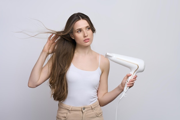 Hermosa mujer joven caucásica con el pelo largo y recto con secador de pelo aislado sobre fondo blanco.