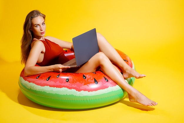 Hermosa mujer joven caucásica en traje de baño aislado sobre fondo amarillo. Concepto de descanso de vacaciones de verano de personas. Siéntese en un anillo inflable de natación, trabaje en una computadora portátil.