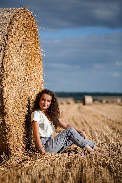 Hermosa mujer joven cerca de un haz de heno en un campo. Vacaciones en el pueblo, una niña disfrutando de la naturaleza en un campo segado en un día soleado