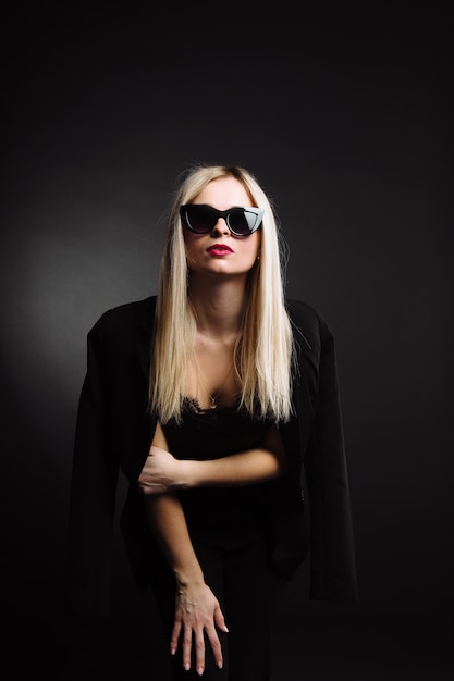 Hermosa mujer joven en chaqueta negra y gafas de sol sobre fondo negro. Bastante rubia chica