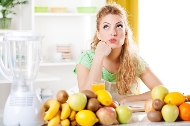 Hermosa mujer joven en una cocina con frutas, pensando. Mirando a la cámara.