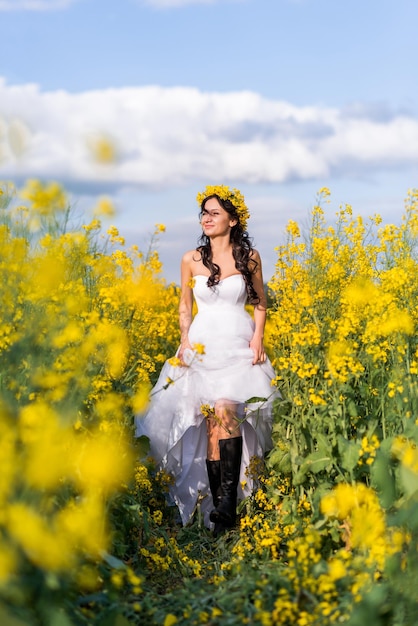 Hermosa mujer joven corre a través de un campo con flores amarillas La novia en un largo vestido blanco y botas en un campo de colza