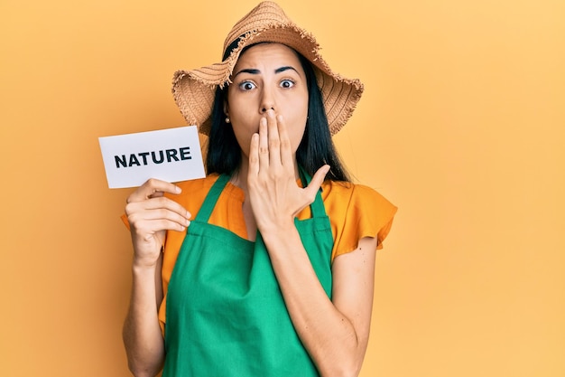 Hermosa mujer joven con delantal de jardinero sosteniendo la palabra de la naturaleza cubriendo la boca con la mano, sorprendida y asustada por error. expresión sorprendida