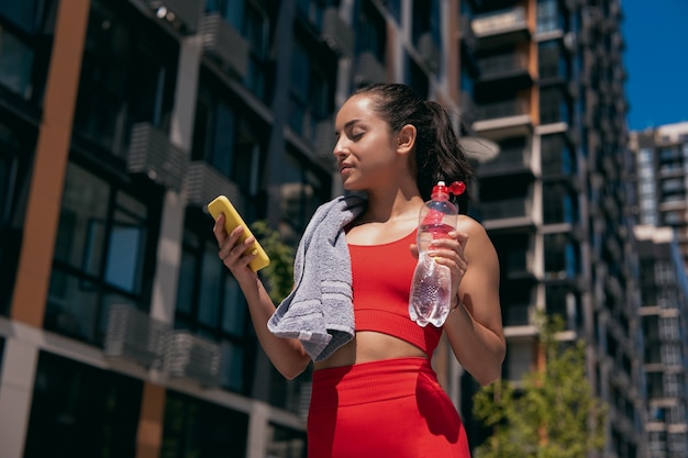 Hermosa mujer joven deportiva con cabello castaño con top rojo y leggins tomando un descanso después de hacer ejercicio o trotar