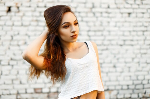 Hermosa mujer joven en una elegante camiseta blanca posando junto a una pared blanca vintage de ladrillo