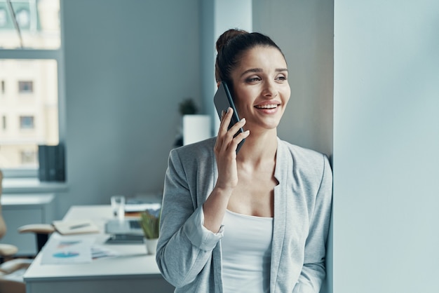 Hermosa mujer joven en elegante casual hablando por teléfono y sonriendo mientras está de pie en la oficina