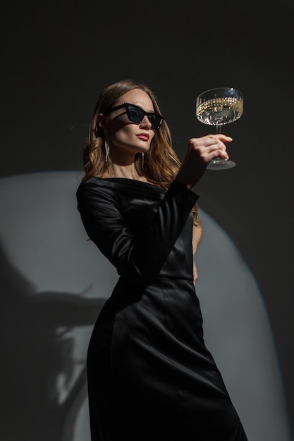 Hermosa mujer joven y elegante con gafas de sol frescas en un elegante vestido negro elegante con una copa de champán vintage sobre un fondo oscuro en una fiesta La dama bonita celebra un evento
