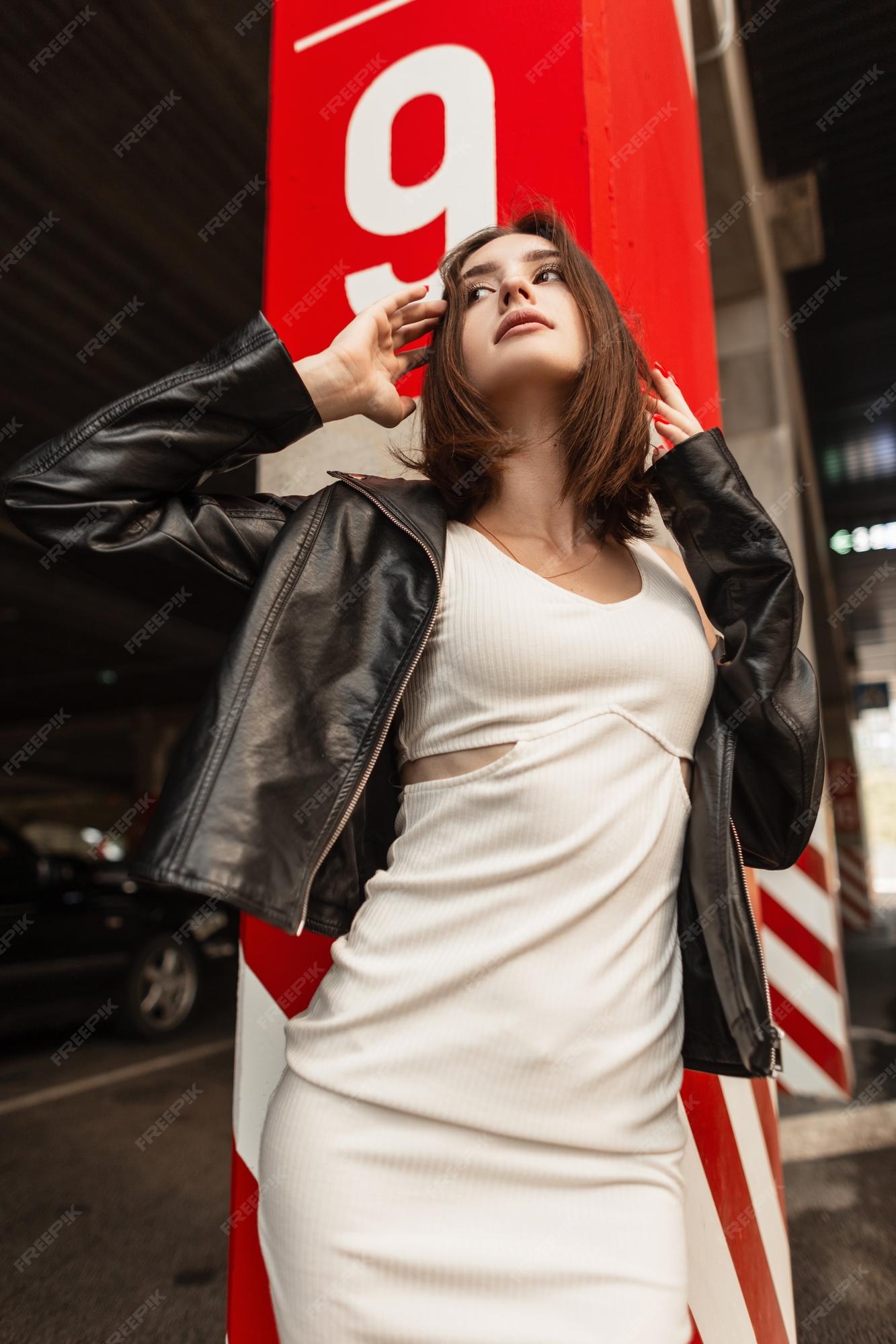 Hermosa mujer joven y un peinado corto en una chaqueta de cuero negro de moda y un vestido blanco posa una columna de hormigón con marcas rojas en