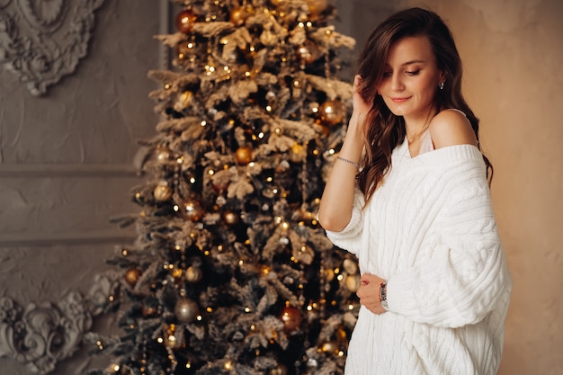 Hermosa mujer joven en elegante vestido blanco posa para la cámara cerca de la Navidad decorada en casa