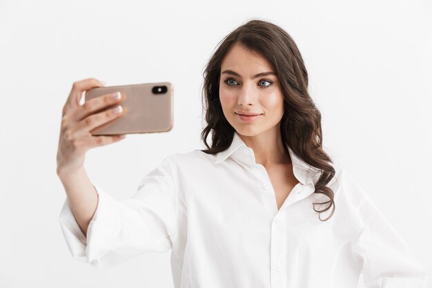 Hermosa mujer joven emocionada con cabello largo y rizado morena con camisa blanca que se encuentran aisladas sobre la pared blanca, tomando un selfie