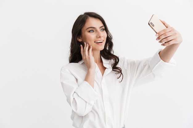 Hermosa mujer joven emocionada con cabello largo y rizado morena con camisa blanca que se encuentran aisladas sobre la pared blanca, tomando un selfie