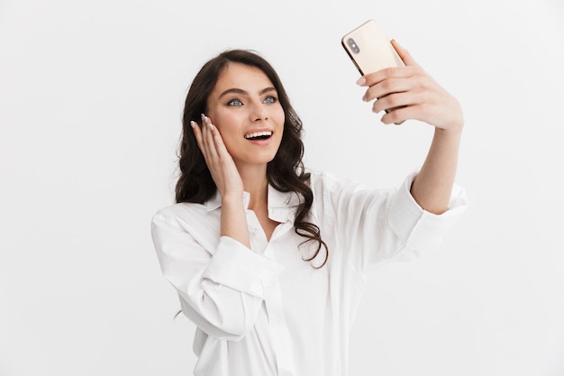 Hermosa mujer joven emocionada con cabello largo y rizado morena con camisa blanca que se encuentran aisladas sobre la pared blanca, tomando un selfie