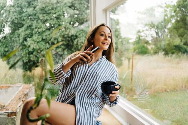 Foto hermosa mujer joven emplazamiento en la encimera de la cocina y navegar en el teléfono móvil i cocina moderna