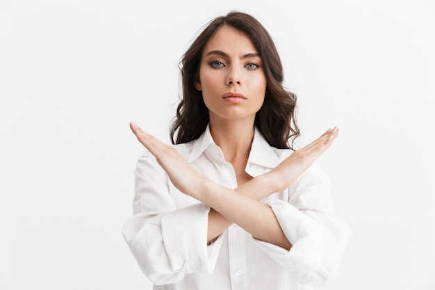 Foto hermosa mujer joven enojada con pelo largo y rizado morena con camisa blanca que se encuentran aisladas sobre una pared blanca, mostrando el gesto de parada