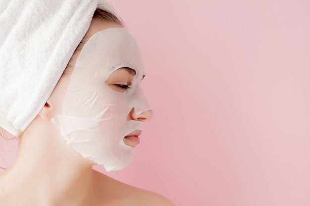 Hermosa mujer joven está aplicando una máscara de tejido cosmético en una cara en una pared rosa.