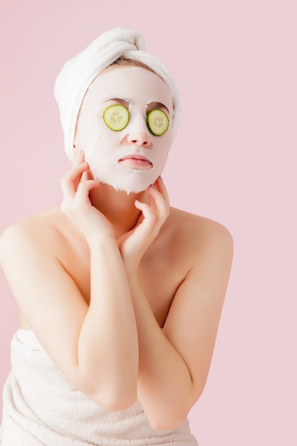 Hermosa mujer joven está aplicando una máscara de tejido cosmético en una cara con pepino en una rosa.