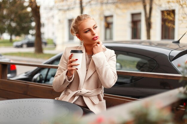 Hermosa mujer joven con estilo en un abrigo se sienta en un café y bebe una bebida caliente al aire libre