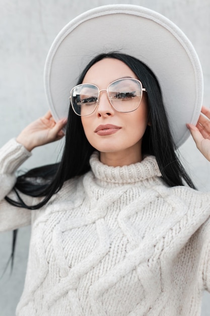 Hermosa mujer joven con estilo con gafas en un suéter de moda vintage y sombrero caminando en la ciudad