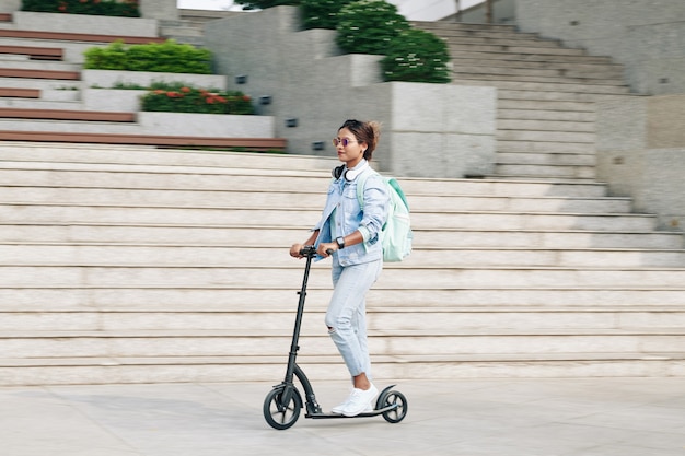 Hermosa mujer joven con estilo con mochila montando en scooter eléctrico en el campus universitario, movimiento borroso