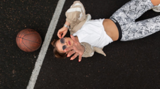 Hermosa mujer joven con estilo en ropa de moda con una pelota tumbada y relajada en la vista del pavimento desde arriba Chica con gafas de sol en ropa deportiva alcanza la cámara