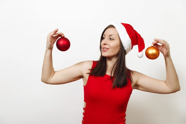 Hermosa mujer joven europea feliz con piel sana y sonrisa encantadora en vestido rojo y sombrero de Navidad con dos juguetes de árbol sobre fondo blanco. Niña de Santa aislada. Concepto de vacaciones de año nuevo 2018.