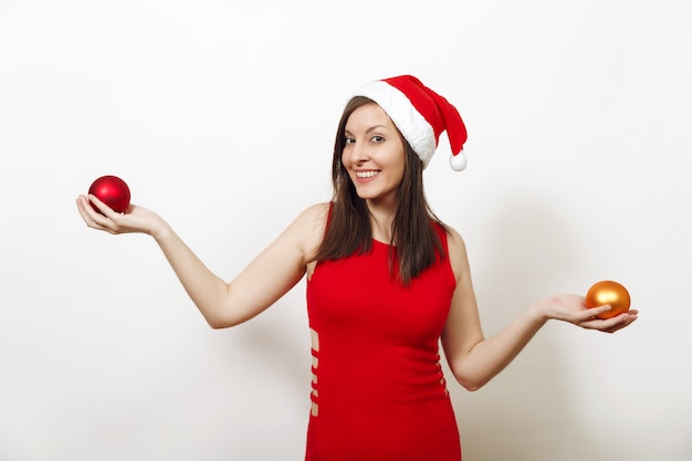 Hermosa mujer joven europea feliz con piel sana y sonrisa encantadora en vestido rojo y sombrero de Navidad con dos juguetes de árbol sobre fondo blanco. Niña de Santa aislada. Concepto de vacaciones de año nuevo 2018.