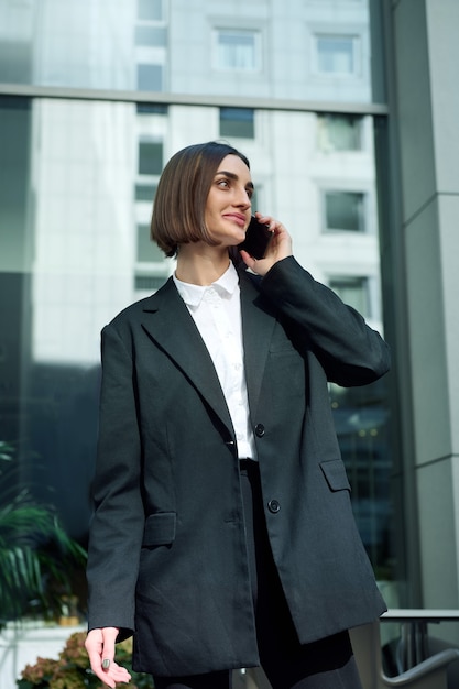 Hermosa mujer joven exitosa segura de sí misma, autónoma, emprendedora, redactora hablando por teléfono celular en el fondo de edificios de gran altura corporativos de oficina moderna. Negocios, concepto de comunicaciones