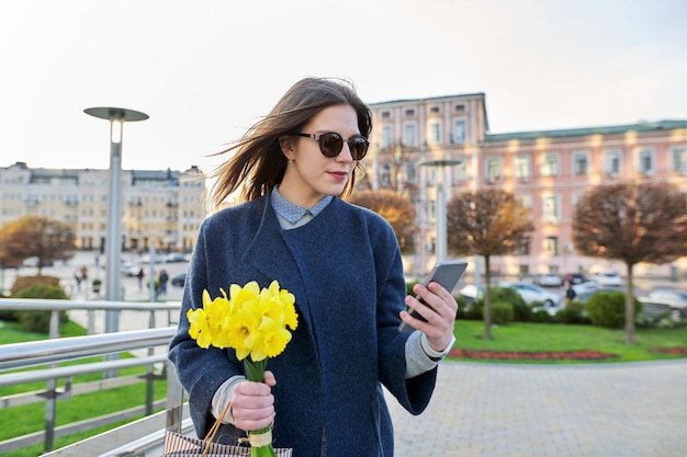 Hermosa mujer joven feliz en abrigo con teléfono con ramo de flores en la calle de la ciudad mujer sonriente mira en la pantalla del teléfono inteligente