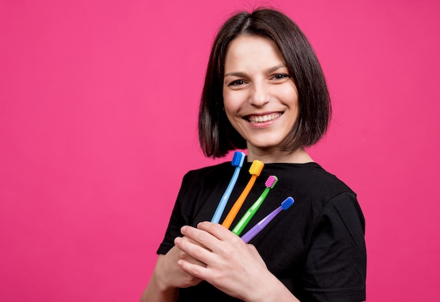 Hermosa mujer joven feliz con cepillos de dientes de diferentes colores sobre fondo rosa en blanco