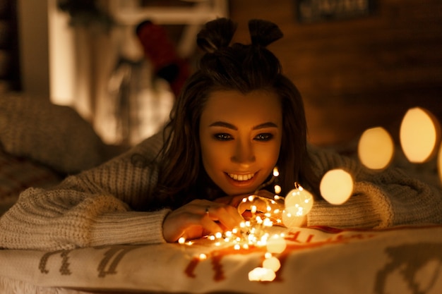 Hermosa mujer joven feliz con luces navideñas en un suéter vintage tejido en la cama