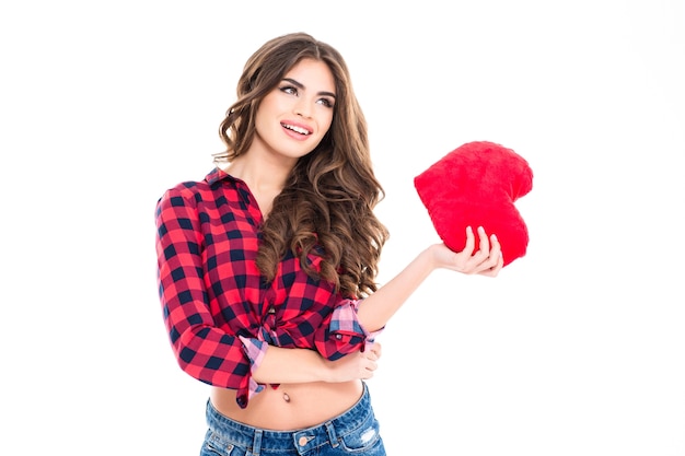 Hermosa mujer joven feliz con el pelo largo y rizado con corazón rojo en la mano de pie sobre la pared blanca